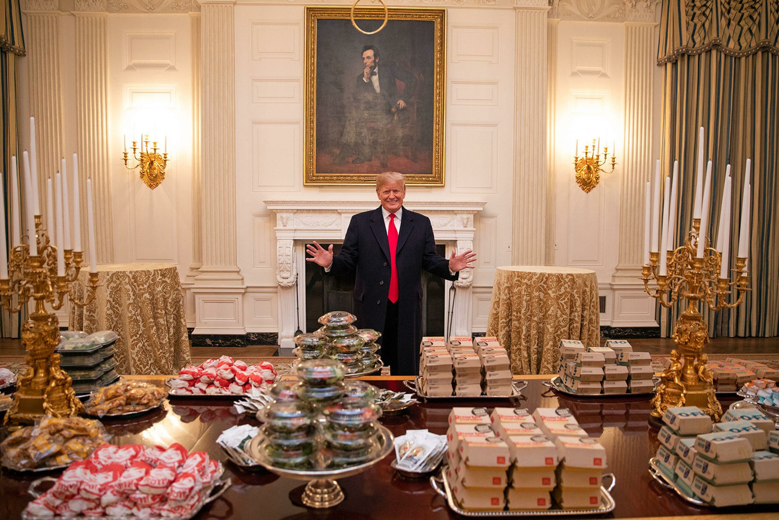 Donald Trump, standing in front of a portrait of Abraham Lincoln, with his arms open, presenting a massive, assorted amount of McDonald's fast food on a table in front of him.